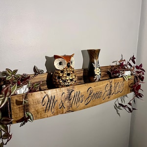 Pu&ograve; includere: Un ripiano in legno curvo con la scritta "Mr. & Mrs. Booze Est. 2022". Il ripiano &egrave; decorato con una statuetta di gufo marrone, un piccolo vaso con motivi floreali e due piante in vaso con viti rampicanti.