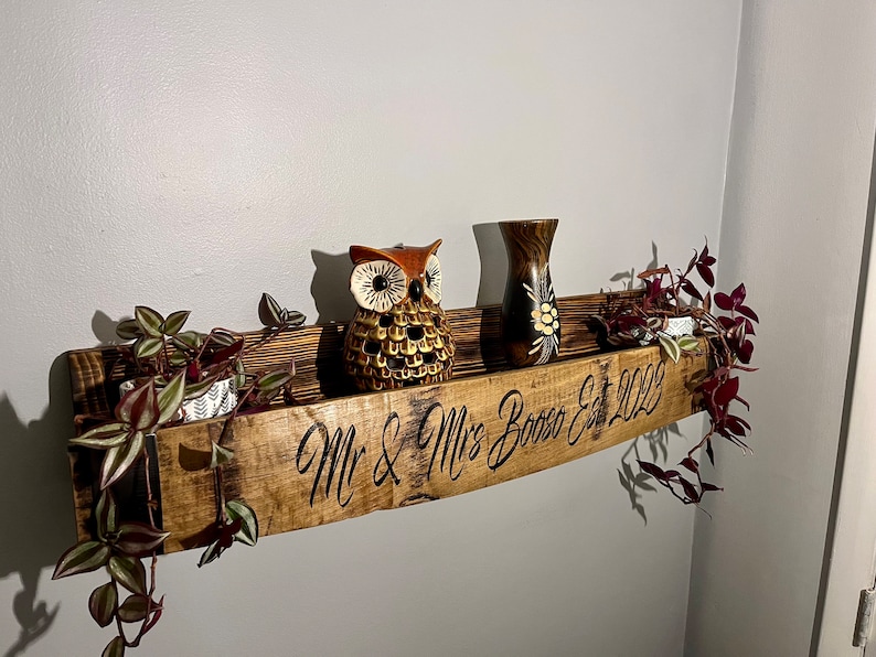 Pu&ograve; includere: Un ripiano in legno curvo con la scritta "Mr. & Mrs. Booze Est. 2022". Il ripiano &egrave; decorato con una statuetta di gufo marrone, un piccolo vaso con motivi floreali e due piante in vaso con viti rampicanti.