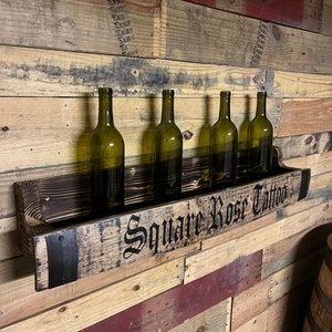 Pu&ograve; includere: Un porta bottiglie di vino in legno con cinque bottiglie di vino. Il porta bottiglie &egrave; fatto di legno riciclato e ha una fascia metallica con il testo "Square Rose Tattoo" su di esso.