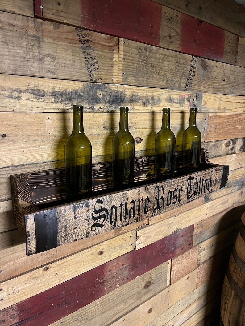 Pu&ograve; includere: Un porta bottiglie di vino in legno con cinque bottiglie di vino. Il porta bottiglie &egrave; fatto di legno riciclato e ha una fascia metallica con il testo "Square Rose Tattoo" su di esso.