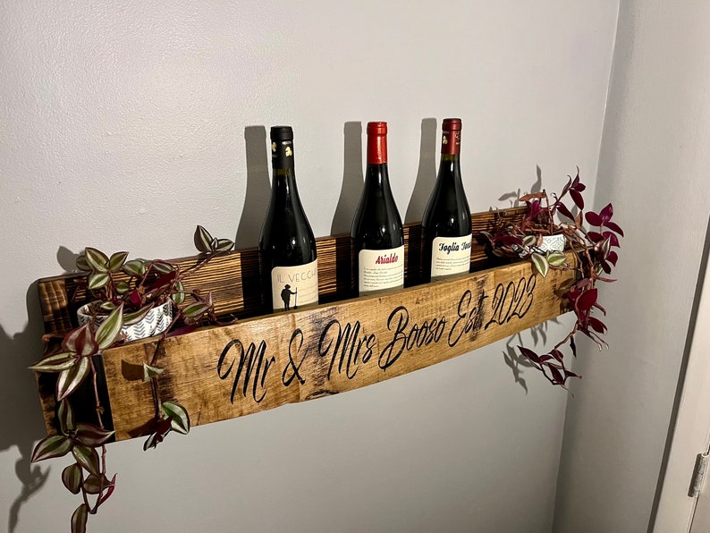 Pu&ograve; includere: Un porta bottiglie in legno con l'iscrizione "Mr & Mrs Bosso Est 2022" e tre bottiglie di vino. Il porta bottiglie &egrave; decorato con del verde.