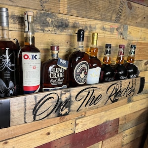 Pu&ograve; includere: Uno scaffale in legno con le parole "On The Rocks" intagliate su di esso. Lo scaffale contiene sette bottiglie di liquore, tra cui Eagle Rare, O.K.I., Elijah Craig e Michters. Lo scaffale &egrave; montato su una parete in legno.
