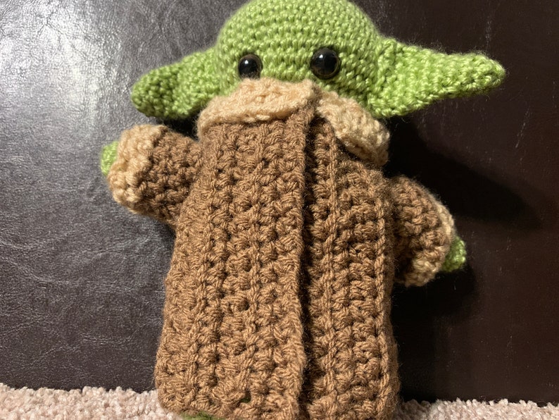 Peut inclure: Un jouet en peluche vert et marron crochet&eacute; ressemblant &agrave; un personnage de la franchise Star Wars. Le jouet porte un manteau marron avec un col beige.