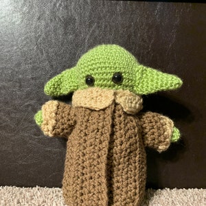Peut inclure: Un jouet en peluche crochet&eacute; repr&eacute;sentant un personnage extraterrestre vert portant un manteau marron. Le jouet a de grands yeux noirs et une petite bouche.
