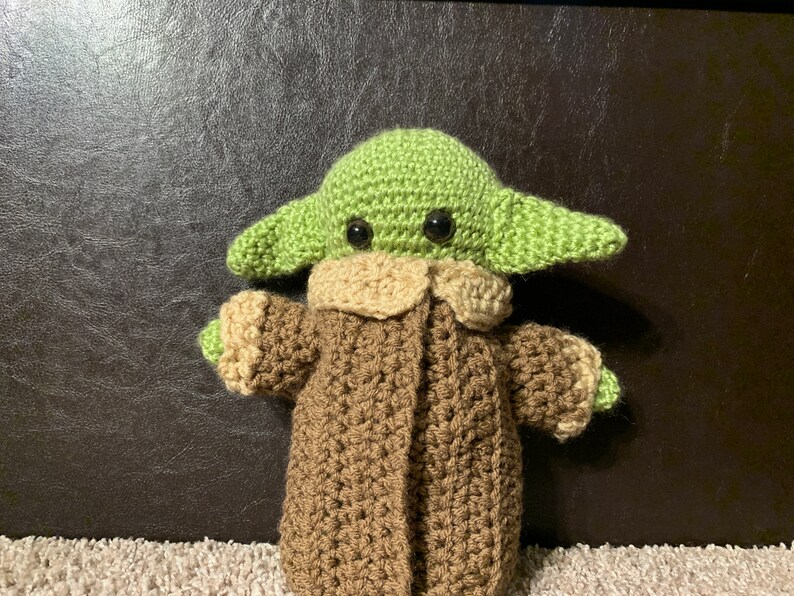 Peut inclure: Un jouet en peluche crochet&eacute; repr&eacute;sentant un personnage extraterrestre vert portant un manteau marron. Le jouet a de grands yeux noirs et une petite bouche.