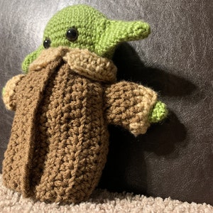 Peut inclure: Un jouet en peluche en crochet repr&eacute;sentant un personnage extraterrestre vert portant un manteau marron. Le jouet a de grands yeux noirs et une petite bouche.