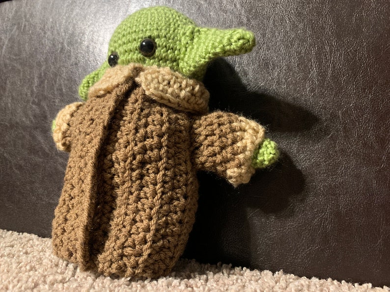 Peut inclure: Un jouet en peluche en crochet repr&eacute;sentant un personnage extraterrestre vert portant un manteau marron. Le jouet a de grands yeux noirs et une petite bouche.