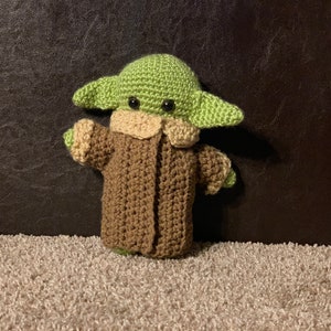 Peut inclure: Un jouet en peluche crochet&eacute; repr&eacute;sentant un personnage vert avec de grandes oreilles, portant un manteau marron. Le jouet a des yeux noirs et une petite bouche.