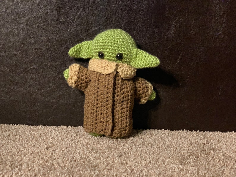 Peut inclure: Un jouet en peluche crochet&eacute; repr&eacute;sentant un personnage vert avec de grandes oreilles, portant un manteau marron. Le jouet a des yeux noirs et une petite bouche.