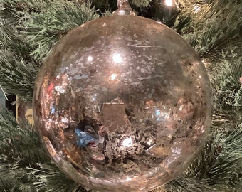 May include: A large, round, silver Christmas ornament with a reflective surface hangs from a silver string. The ornament is set against a backdrop of green Christmas tree branches and small white lights, creating a festive holiday scene.