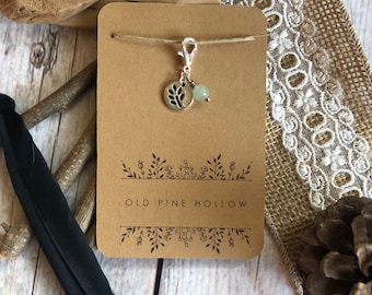 May include: A silver charm with a tree design and a green bead hangs from a brown card with the text "OLD PINE HOLLOW" in black lettering.
