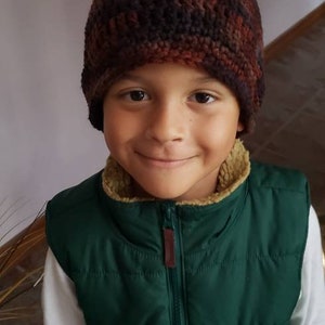 May include: A young person wearing a brown and black crocheted hat and a green puffer vest. The vest has a zipper and a brown zipper pull.