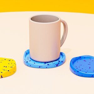 May include: Three coasters with a speckled design. One coaster is yellow with blue speckles, one is blue with purple speckles, and one is blue with purple speckles. A light gray mug sits on top of the blue coaster.
