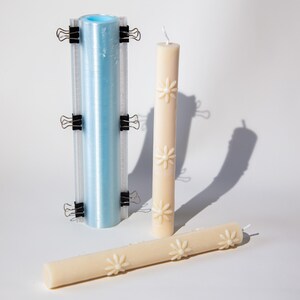 May include: A blue plastic tube mould with black binder clips and a white candle with a floral design. The candle is lying on its side.