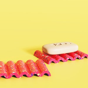 Puede incluir: Dos jaboneras onduladas con manchas de color rosa y blanco. Una jabonera tiene una pastilla de jab&oacute;n blanca encima.