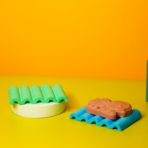 Puede incluir: Tres moldes de silicona de colores sobre una superficie amarilla. Los moldes tienen forma de l&iacute;neas onduladas y son verdes, azules y turquesa. Un molde tiene un objeto de silicona marr&oacute;n en su interior.
