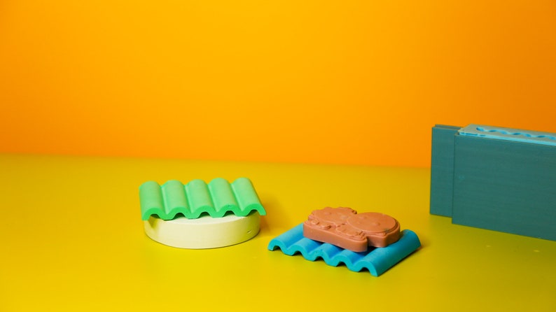 Puede incluir: Tres moldes de silicona de colores sobre una superficie amarilla. Los moldes tienen forma de l&iacute;neas onduladas y son verdes, azules y turquesa. Un molde tiene un objeto de silicona marr&oacute;n en su interior.