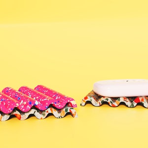 Puede incluir: Dos jaboneras hechas de materiales reciclados. La jabonera de la izquierda es rosa con motas blancas y azules. La jabonera de la derecha es verde, marr&oacute;n y roja con motas blancas. Una pastilla de jab&oacute;n blanca est&aacute; sobre la jabonera verde.