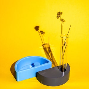 May include: A blue and gray ceramic vase with a single dried flower stem in a clear glass tube. The vase is on a yellow background.