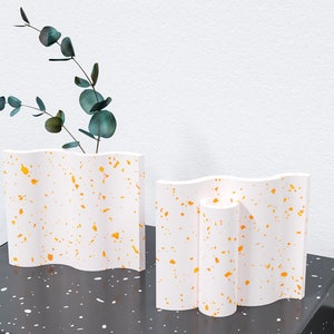May include: Two white ceramic vases with a wavy design and orange speckles. The vase on the left has a sprig of green eucalyptus leaves.