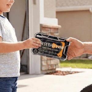 May include: A black and orange tool kit with the text "BEST DAD EVER" is being handed from a young boy to a man. The man is sitting next to a bicycle. The tool kit has the words "Love, Lots & Giggles" on it.