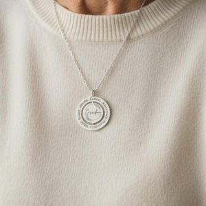 May include: A silver pendant necklace with a circular charm. The charm has engraved text around the edge and a central word. The woman in the image is wearing a cream-colored sweater.