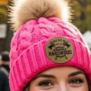 May include: A bright pink knit beanie with a large faux fur pom-pom on top. The hat features a circular leather-like patch on the front with the text "CHICKS DIG MEN HANDLE HARDWOOD" surrounding a graphic of two crossed axes in black ink.