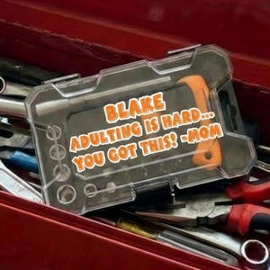 May include: A red metal toolbox filled with various tools, including a wrench, hammer, and pliers. A clear plastic case with an orange tool is visible, with the text "BLAKE ADULTING IS HARD... YOU GOT THIS! -MOM".