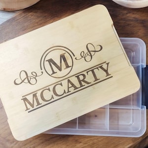 May include: A personalized bamboo cutting board with a clear plastic storage container underneath. The cutting board has a monogrammed initial "M" in a circle with the name "MCCARTY" engraved below.