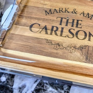 May include: A wooden cutting board with a clear plastic lid. The board is engraved with the text "Mark & Amy The Carlsons" and a floral design.
