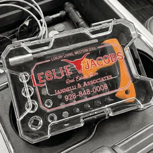 May include: A clear plastic tool kit with a black interior, containing various bits and an orange handle. The kit features the text "LESLIE JACOBS Real Estate Team" and a phone number. The kit is in a car console.