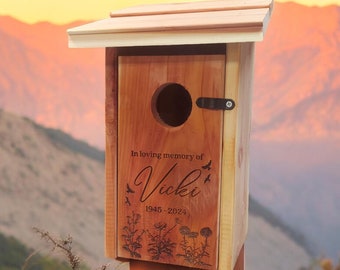 Puede incluir: Una casita para pájaros de madera con un tejado inclinado y un agujero de entrada circular. La casita está grabada con el texto "In loving memory of Vicki 1945-2024" junto con ilustraciones florales y de pájaros. Está montada en un poste de madera.