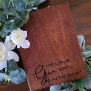 May include: A wooden cutting board with the text "It is always Gin o'clock at the Engelmann's" engraved on it. The board is a dark reddish brown color and has a natural wood grain.