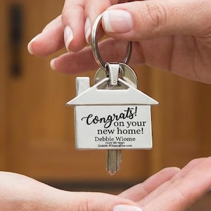 Op de afbeelding: Een zilveren sleutelhanger in de vorm van een huis met de tekst "Congrats! on your new home!" wordt overhandigd. De sleutel is bevestigd aan een zilveren ring. De achtergrond is een wazige afbeelding van een houten deur.