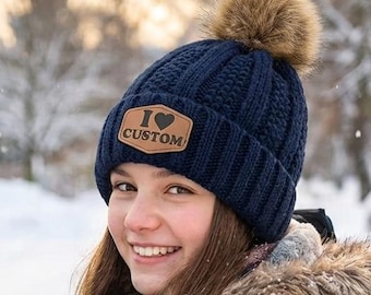May include: A navy blue knit beanie with a brown faux fur pom-pom on top. The beanie features a brown leather-like patch on the front with the text I heart CUSTOM, where the heart is represented by a symbol. The hat has a ribbed texture.