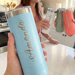 May include: A light blue insulated tumbler with the name "Amanda" in a cursive font. The tumbler has a silver lid and is held in front of a kitchen setting. Other tumblers in white, green, and coral are also visible.