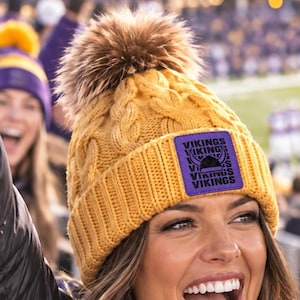 May include: A golden yellow cable-knit beanie with a brown faux fur pom-pom on top. The beanie features a square purple patch on the front with a black graphic of a helmet and the repeated text "VIKINGS" in black lettering.
