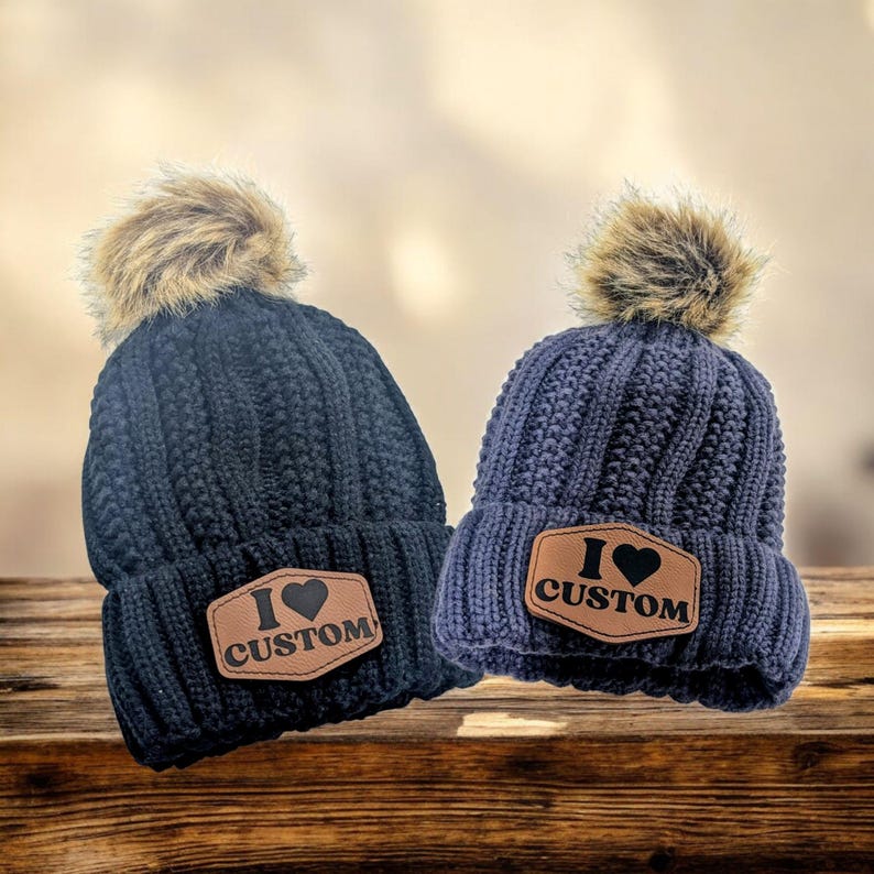 Puede incluir: Dos gorros de punto azul marino con pompones de piel sint&eacute;tica. Cada gorro tiene un parche de cuero marr&oacute;n que dice "I &hearts; CUSTOM". Los gorros se exhiben sobre una superficie de madera.