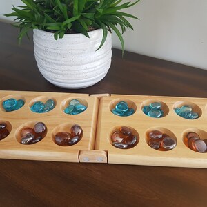 May include: A wooden Mancala board game with two rows of six playing pits and two larger storage pits. The pits are filled with blue and brown glass stones.