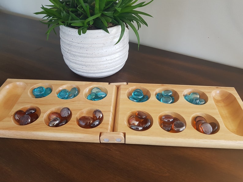 May include: A wooden Mancala board game with two rows of six playing pits and two larger storage pits. The pits are filled with blue and brown glass stones.