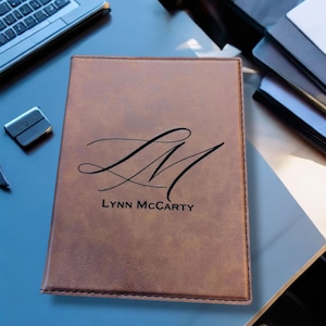 May include: A brown leather journal with a stitched edge and a personalized monogram of "LM" with the name "Lynn McCarty" engraved below.
