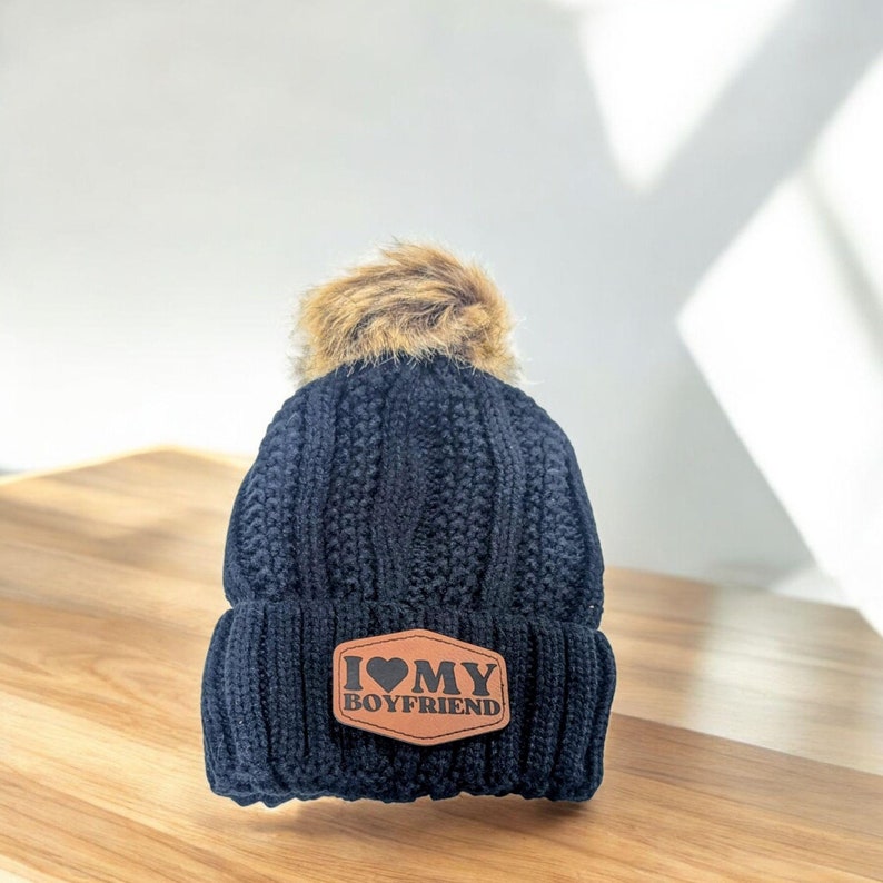 Puede incluir: Un gorro de punto trenzado en color azul marino oscuro con un pomp&oacute;n de piel sint&eacute;tica beige en la parte superior. El dobladillo frontal muestra un parche hexagonal marr&oacute;n de aspecto cuero con el texto I HEART MY BOYFRIEND.