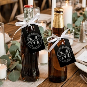 Op de afbeelding: Twee glazen flessen, één doorzichtig en één amberkleurig, elk voorzien van een zwart rechthoekig flesopener-label vastgebonden met een wit satijnen lint. De labels zijn bedrukt met een cirkelvormig monogramlogo met de tekst Taylor & Mackenzie, Metcalf, Est. 2024.