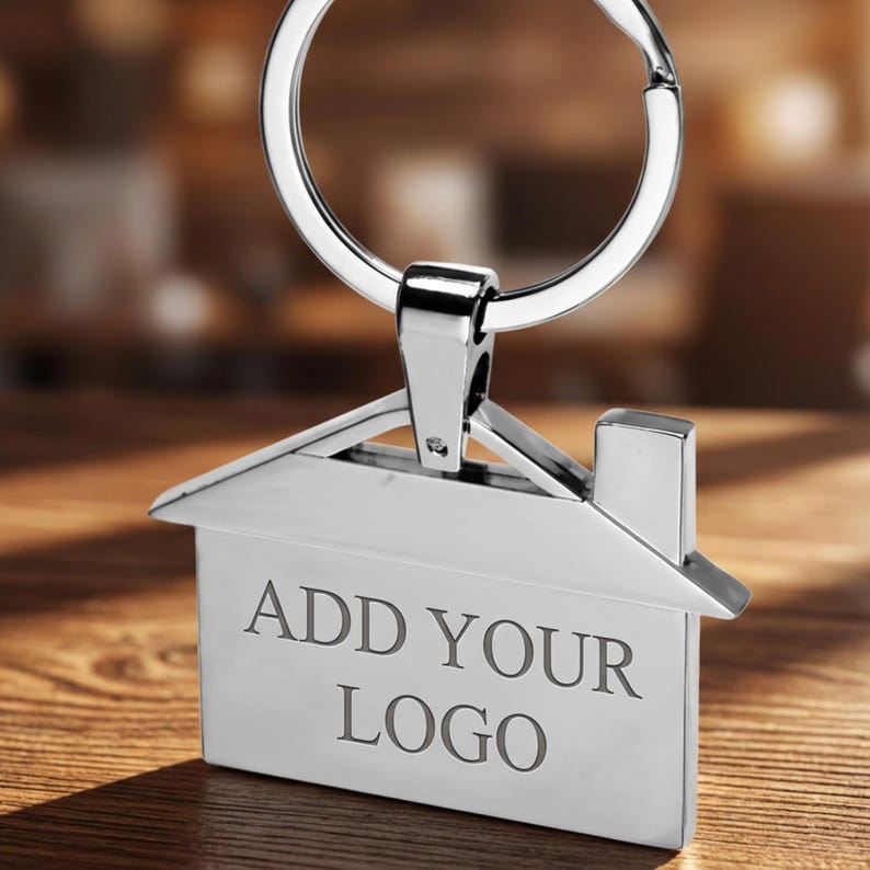 Op de afbeelding: Een zilverkleurige sleutelhanger in de vorm van een huis met een sleutelring. De sleutelhanger heeft de tekst "ADD YOUR LOGO" gegraveerd op de voorkant. De achtergrond is een vage houten ondergrond.