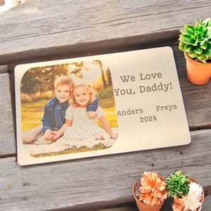 May include: A rectangular metal plaque with rounded corners, featuring a color photograph of two children. The text "We Love You, Daddy!" is printed on the right side, along with names and the year 2024. The plaque is on a wooden surface.