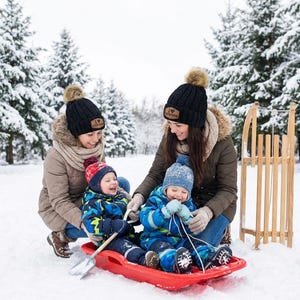 Puede incluir: Dos gorros de punto negro con pompones de piel sint&eacute;tica marr&oacute;n y un parche de cuero beige con el texto I HEART CUSTON. Los gorros los llevan dos adultos en un entorno nevado mientras dos ni&ntilde;os est&aacute;n sentados en un trineo de pl&aacute;stico rojo.