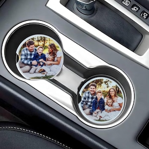 May include: Two round car coasters with a white background and a photo of a family of four. The family is smiling and standing in a grassy area. The coasters are in the cup holders of a car.