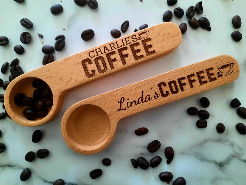 May include: Two wooden coffee scoops with personalized names and the word "Coffee" engraved on them. The scoops are on a white marble surface with coffee beans scattered around them.
