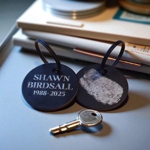 May include: Two black round keychains with metal rings. One keychain has the name "SHAWN BIRDSALL" and the years "1988-2025" engraved in white. The other keychain has a white fingerprint. A gold key rests below.