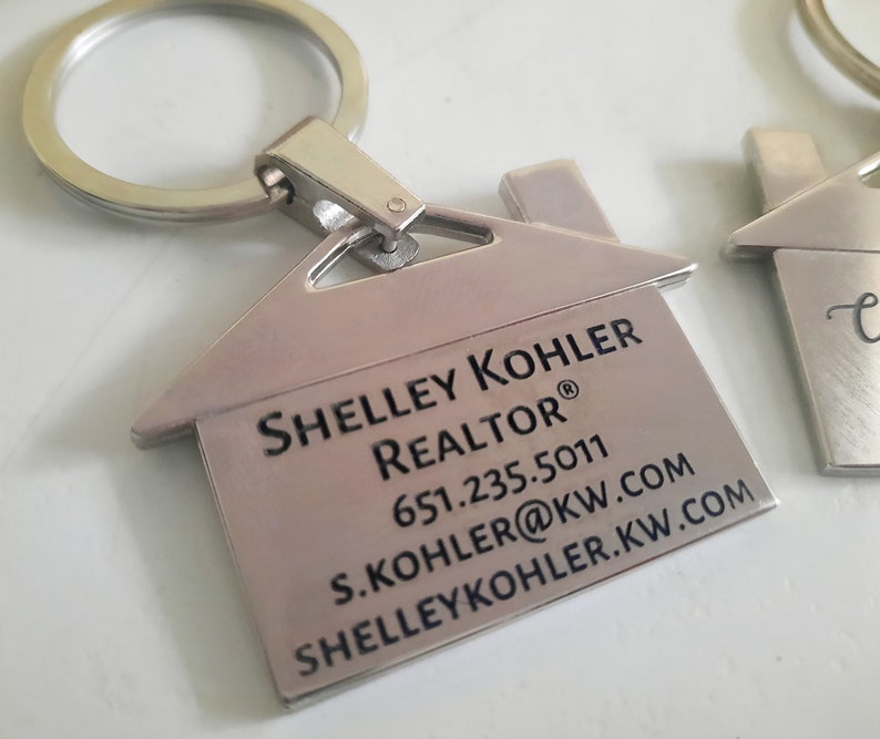 Op de afbeelding: Zilverkleurige sleutelhangers in de vorm van een huis. E&eacute;n sleutelhanger heeft de tekst "Shelley Kohler, Realtor" met contactgegevens. De sleutelhangers hebben een zilveren ring voor sleutels. Eenvoudig, modern ontwerp.